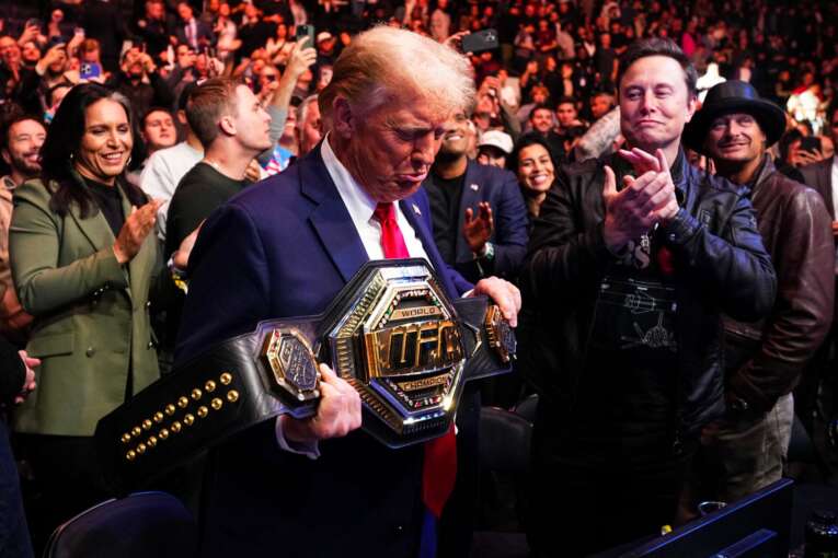 Donald Trump in a suit and red tie holds a wrestling championship belt as a crowd cheers around him.