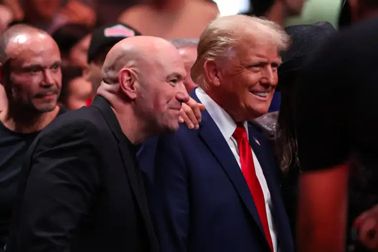 Two men in suits smiling together in a crowded event, one leaning toward the other for a quick greeting.