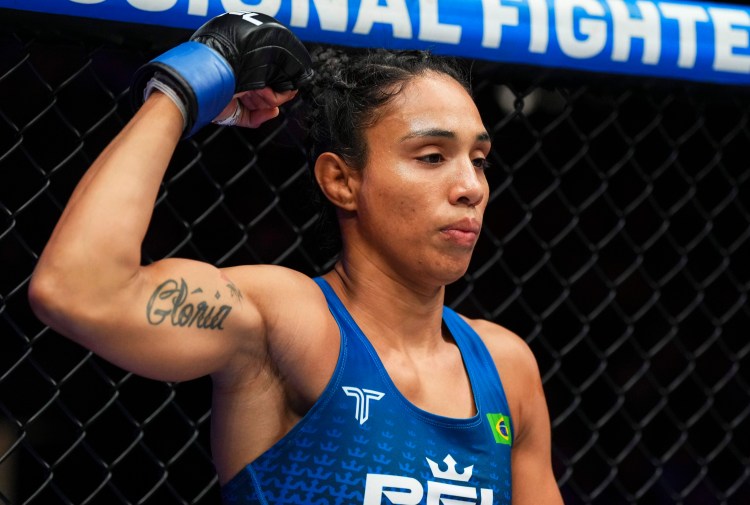 Female mixed martial artist in blue gear raises her arm inside a chain-link cage during a bout, tattoo reading 'Glória' on her left arm.