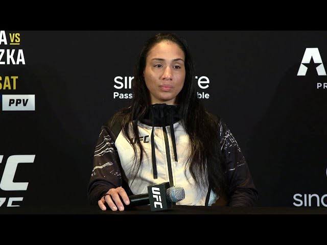 Female mixed martial artist at a UFC press conference, seated at a table with a UFC microphone in front, black backdrop behind her.