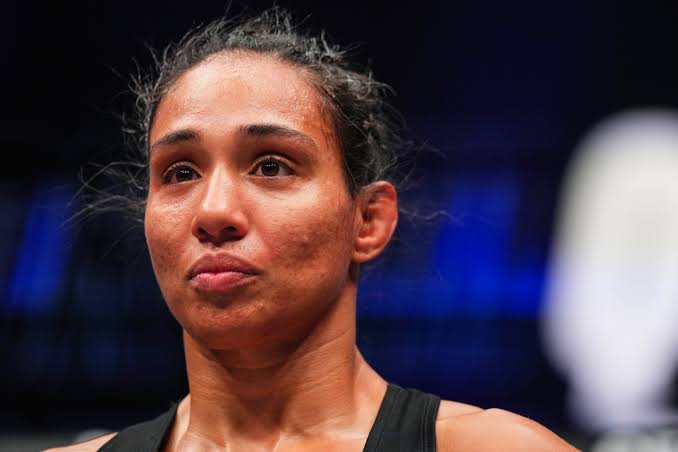 Close-up of a female athlete with dark hair tied back, wearing a black sleeveless top, looking focused in a blue-lit arena.