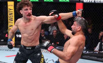 Two UFC fighters exchange punches in the octagon; the left fighter throws a straight, while the right blocks with an arm raised behind his head, crowd watching in the background.