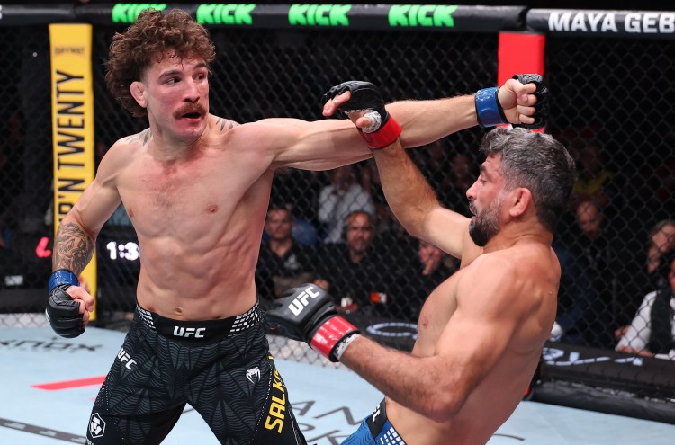 Two UFC fighters exchange punches in the octagon; the left fighter throws a straight, while the right blocks with an arm raised behind his head, crowd watching in the background.