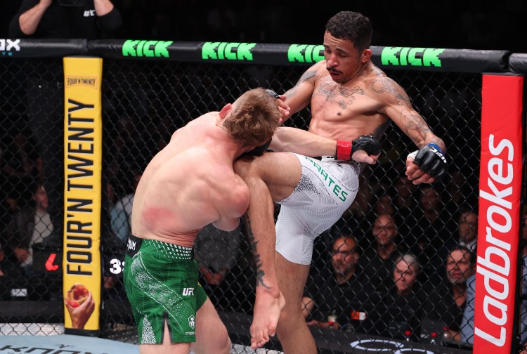 A mixed martial arts bout inside a cage: a fighter in white shorts delivering a high kick to a opponent in green shorts while spectators watch from the sidelines.