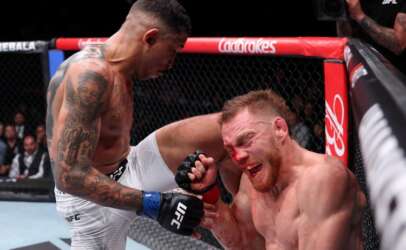 Two male UFC fighters in the octagon; one standing and delivering a knee to the other’s bloody face against the cage.
