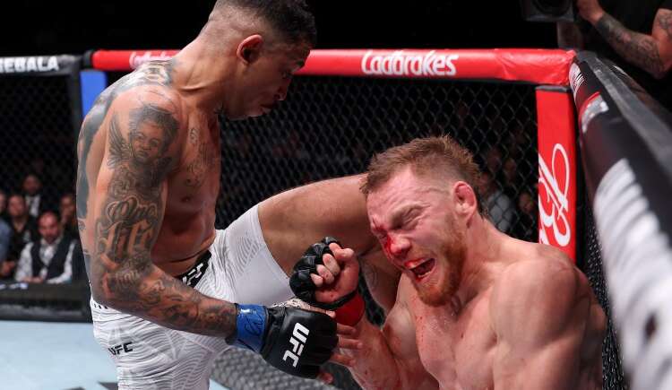 Two male UFC fighters in the octagon; one standing and delivering a knee to the other’s bloody face against the cage.