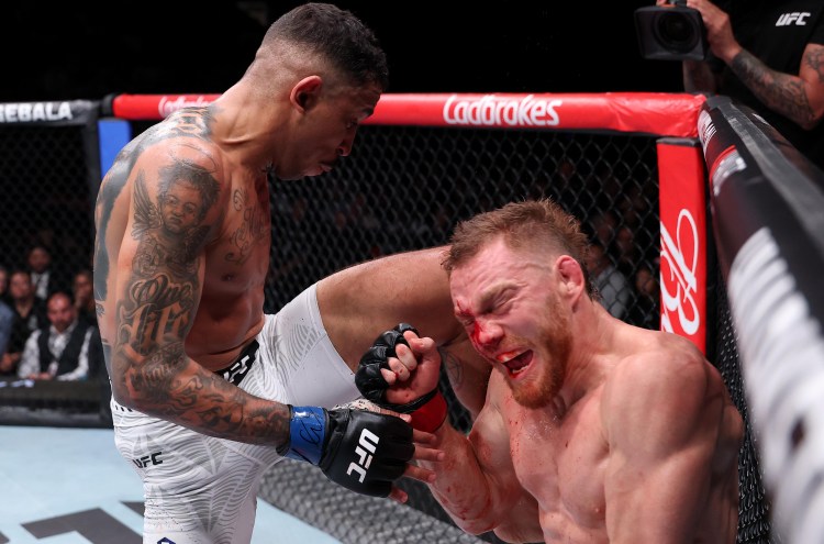 Two male UFC fighters in the octagon; one standing and delivering a knee to the other’s bloody face against the cage.
