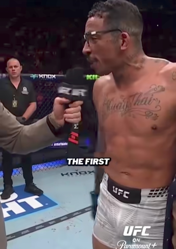 UFC fighter shirtless in white shorts being interviewed inside the octagon, microphone held up by a interviewer, with arena spectators in the background and