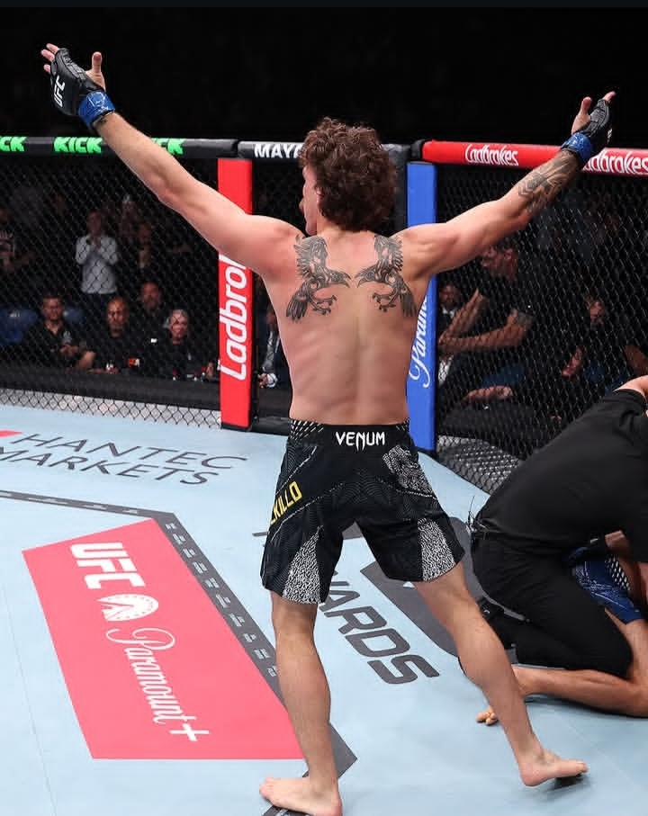 MMA fighter with arms raised in the octagon, back tattoos visible, wearing black shorts with logos, celebrating victory.