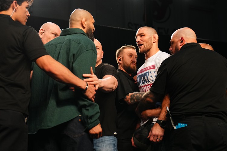 Group of men in a black-clad circle surrounding a bearded fighter in a white 'BREAK' T-shirt on a stage, hands on shoulders as they talk seriously.