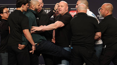 Bald man in a black shirt delivers a high kick while several other men grab or hold him back on a stage with sponsor banners.