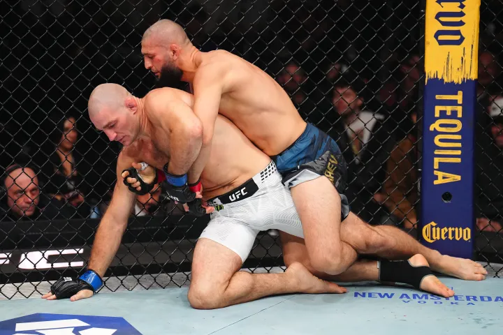 Two male UFC fighters grapple on the mat inside the octagon; the top fighter in blue shorts grips the bottom fighter in white shorts in a body-lock.