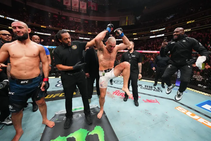 Fighter in the center raises his arms in victory inside the UFC octagon as referees and teammates surround him, crowd watching in the arena.