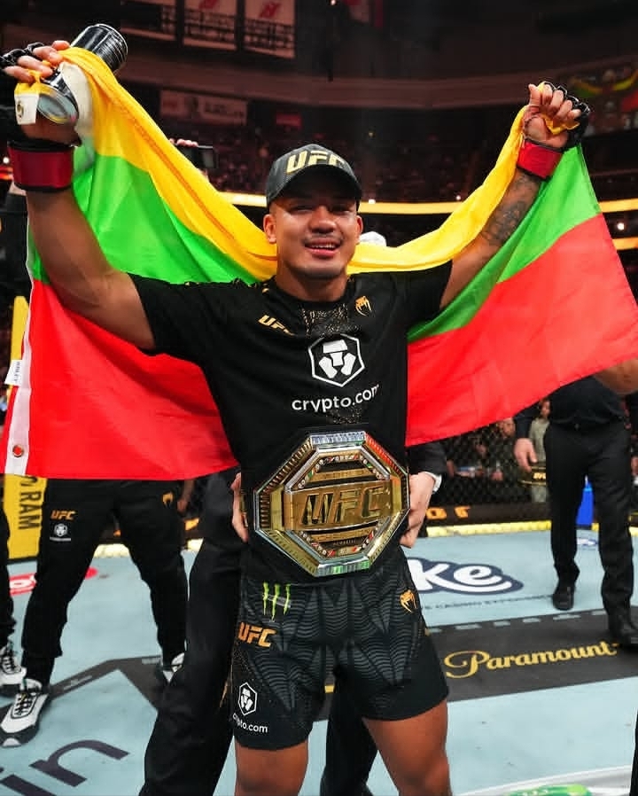 UFC fighter in victory pose holding a bright green-yellow-red flag behind him, wearing a championship belt inside the octagon.
