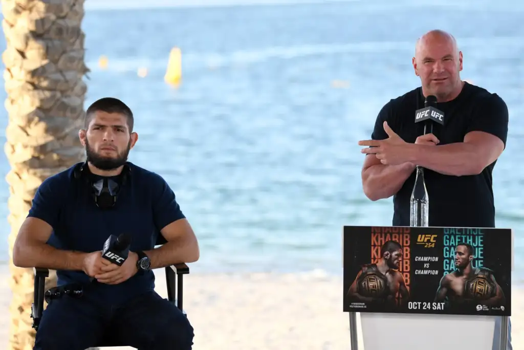 Two UFC figures at a beach press conference; man on the right speaks into a UFC microphone while gesturing, and a fighter sits on the left with gear nearby.