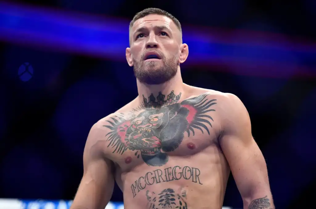 Bearded, tattooed male fighter shirtless in a dim arena, looking upward with blue stage lights in the background.