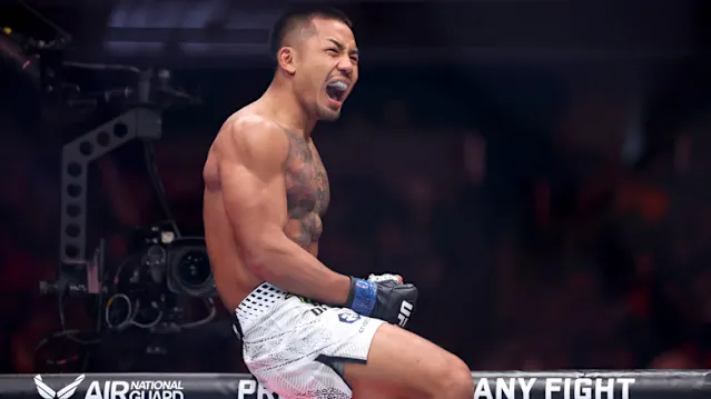 Mixed martial artist shirtless in white shorts, clenched fist, shouting in a fighting arena after a match.