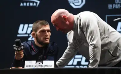 UFC press conference: a fighter sits at a table with a microphone while a bald man in a gray hoodie leans in to speak.