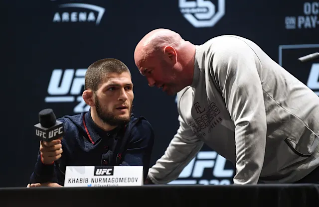 UFC press conference: a fighter sits at a table with a microphone while a bald man in a gray hoodie leans in to speak.