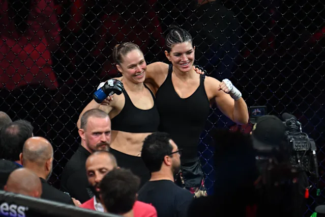 Two female mixed martial artists in black sports bras pose together inside a cage, smiling with arms around each other, surrounded by spectators and a camera crew.