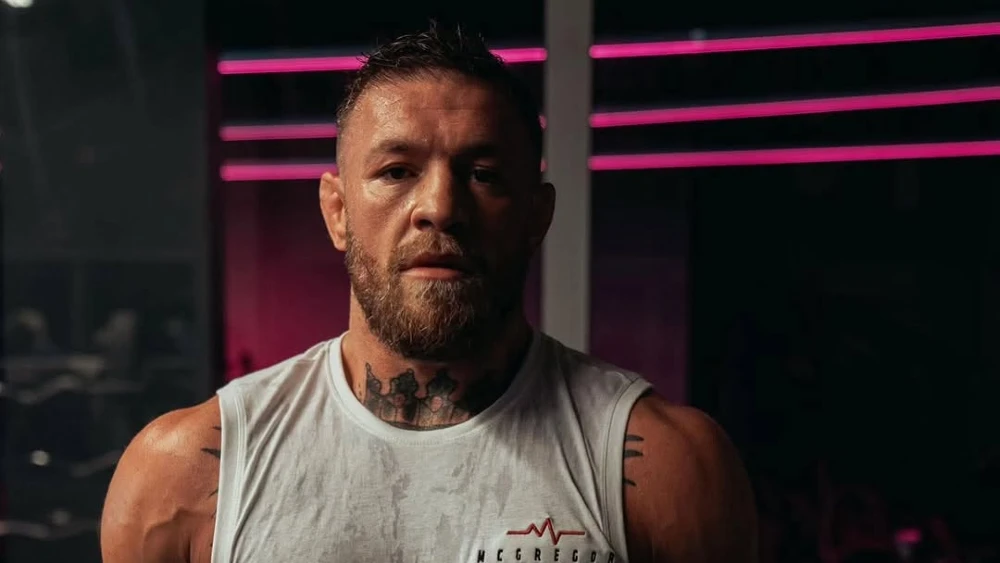 Close-up of a muscular man with a beard wearing a white sleeveless shirt in a dim gym lit by pink neon lights behind him, looking at the camera.
