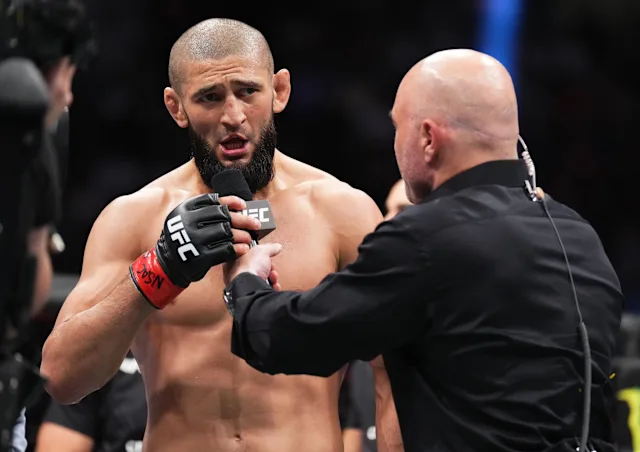 Bearded male mixed martial artist speaks into a microphone during a post-fight interview, a bald interviewer in black holds the mic close.