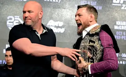 Two men on a stage exchange a handshake; the man on the left is bald in a black polo, the man on the right wears a purple suit and a large championship belt.