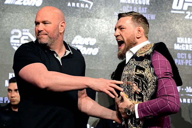 Two men on a stage exchange a handshake; the man on the left is bald in a black polo, the man on the right wears a purple suit and a large championship belt.