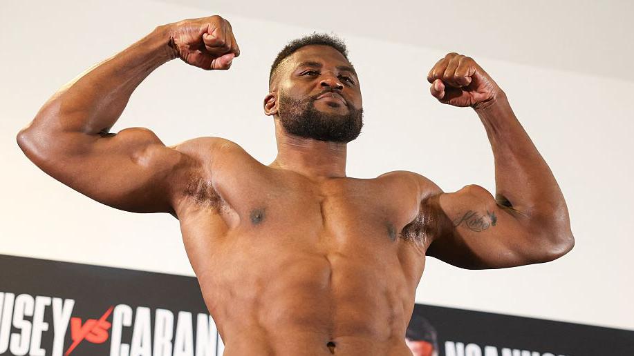 Muscular Black man, shirtless, flexing his biceps on stage with a sports banner in the background.