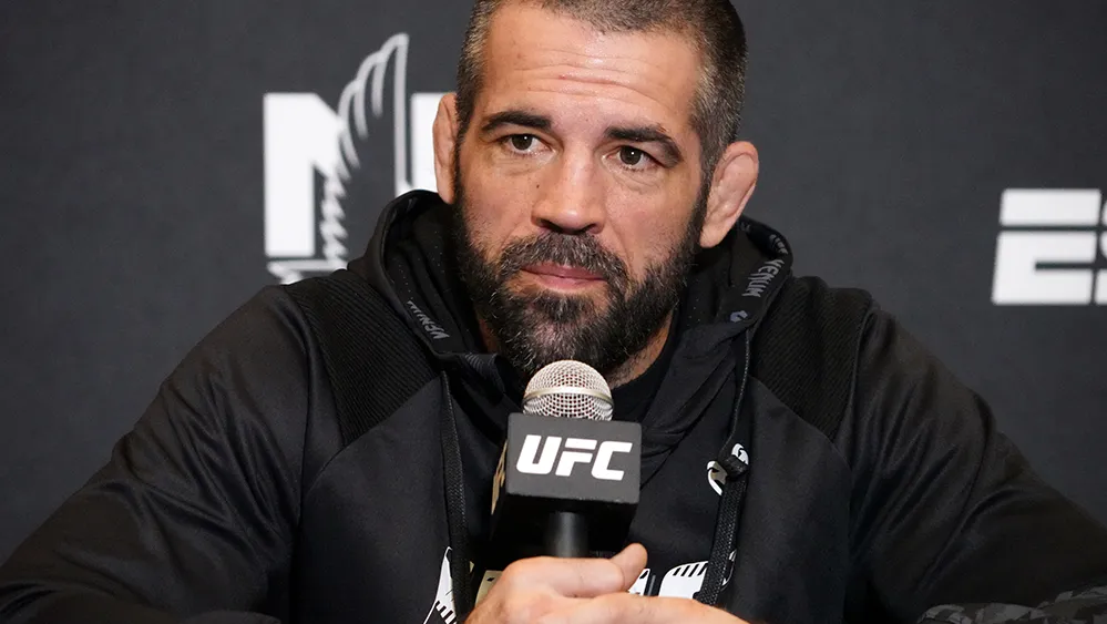 MMA fighter at a UFC press conference, speaking into a UFC-branded microphone, wearing a black hoodie.