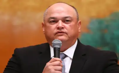 Man in a dark suit speaks into a handheld microphone during a formal event or conference, with a warm orange backdrop behind him.
