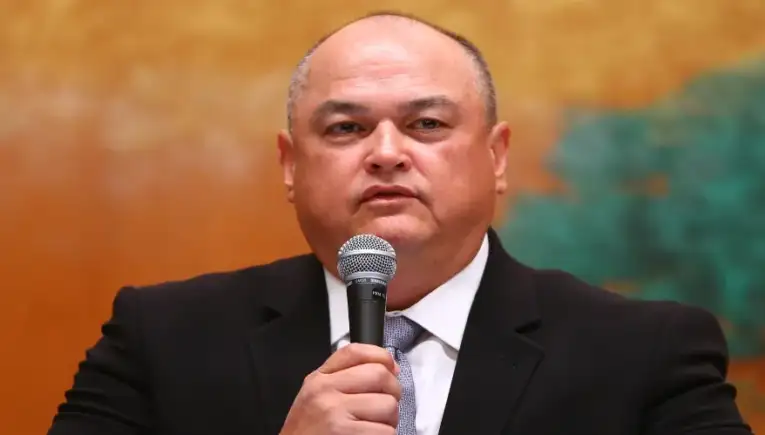 Man in a dark suit speaks into a handheld microphone during a formal event or conference, with a warm orange backdrop behind him.