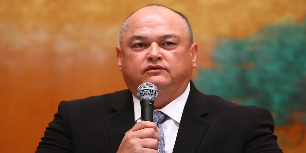 Man in a dark suit speaks into a handheld microphone during a formal event or conference, with a warm orange backdrop behind him.