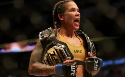 Female UFC fighter celebrating with two championship belts draped over her shoulders, fists raised in victory