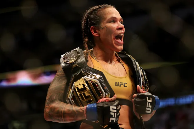Female UFC fighter celebrating with two championship belts draped over her shoulders, fists raised in victory