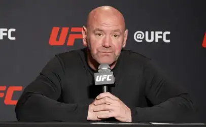 Bald man in a black long-sleeve shirt sits at a press table, hands clasped, speaking into a UFC microphone against a dark backdrop.