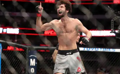 UFC fighter raises a finger in triumph inside the octagon after a win, shirtless with red wristbands and crowd in the background.