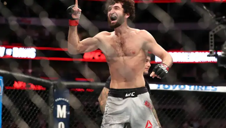 UFC fighter raises a finger in triumph inside the octagon after a win, shirtless with red wristbands and crowd in the background.