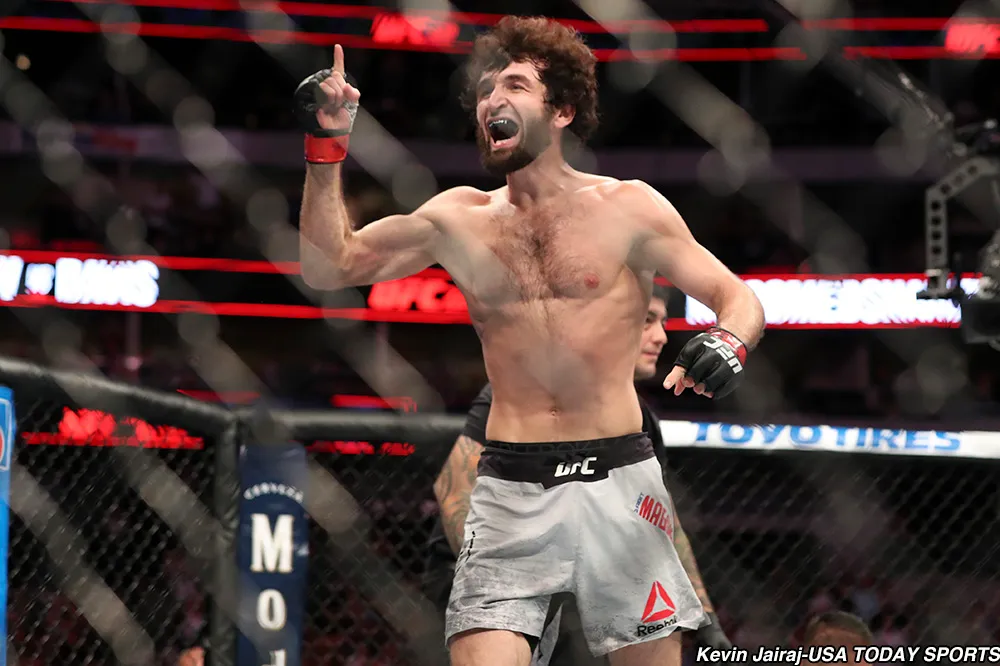 UFC fighter raises a finger in triumph inside the octagon after a win, shirtless with red wristbands and crowd in the background.