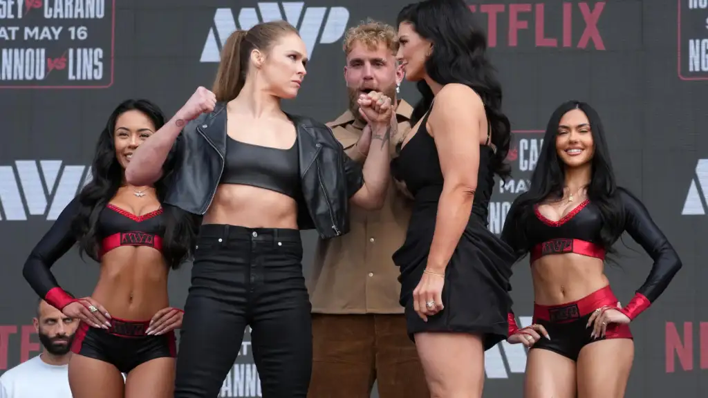Group of six people on a stage with a dark backdrop; two dancers in black and red outfits flank a bearded man and a woman in a black dress who appear to be in conversation.