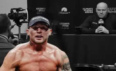 Shirtless MMA fighter wearing a cap, sweating, facing the camera at a press conference with a cameraman and a questioning figure in the background.