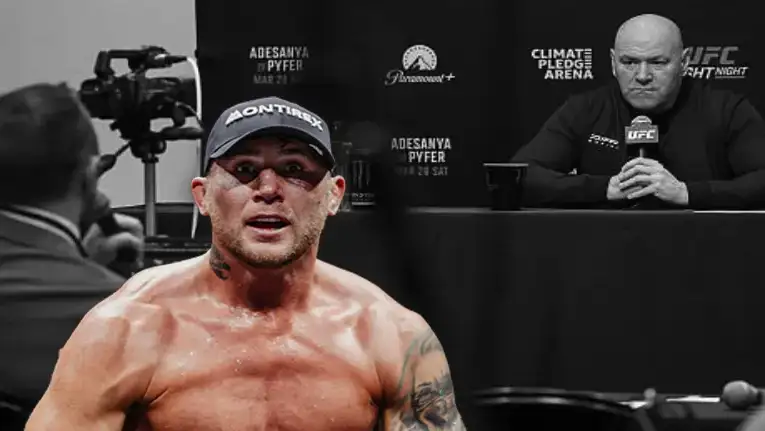 Shirtless MMA fighter wearing a cap, sweating, facing the camera at a press conference with a cameraman and a questioning figure in the background.