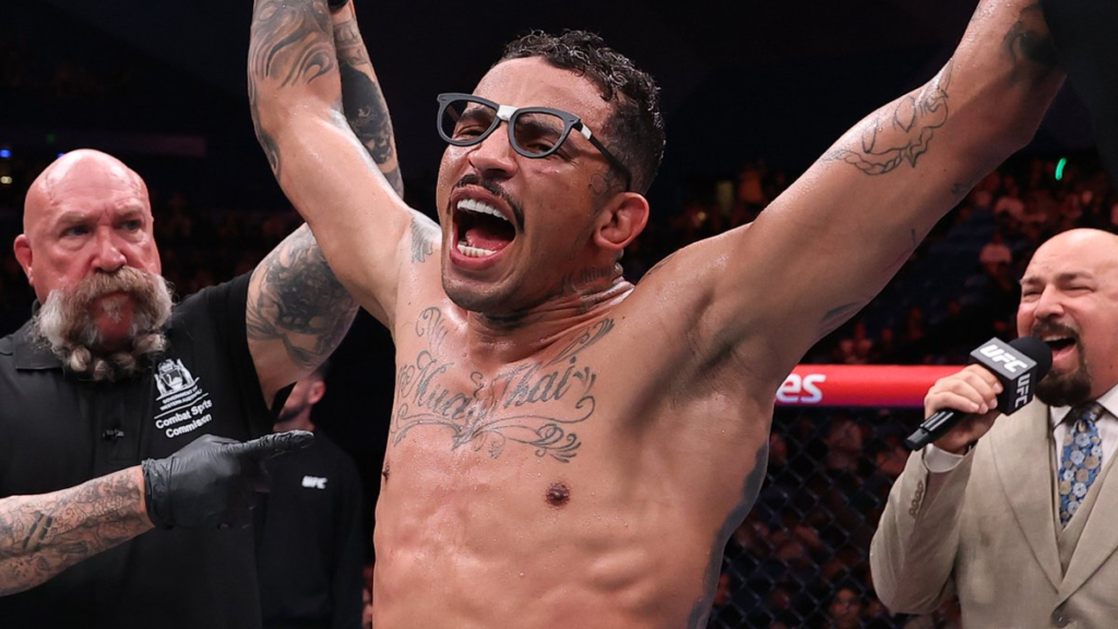 UFC fighter with glasses raises arms in victory inside the octagon, crowd cheering and announcer nearby with mic nearby.