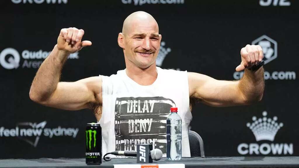 8d2dedf690359bd6a5ead6040314e2c2 - AfroCombat Bald male mixed martial artist flexes his arms and smiles at a press conference, with a Monster energy can and a water bottle on the table in front of him.