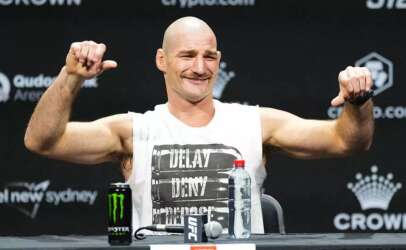 Bald male mixed martial artist flexes his arms and smiles at a press conference, with a Monster energy can and a water bottle on the table in front of him.