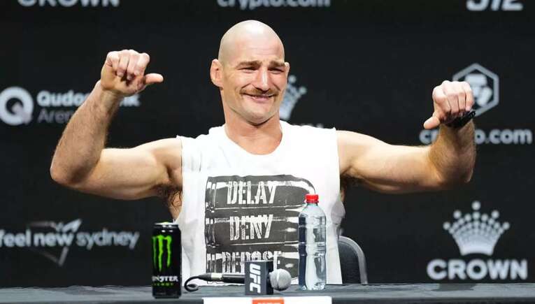Bald male mixed martial artist flexes his arms and smiles at a press conference, with a Monster energy can and a water bottle on the table in front of him.