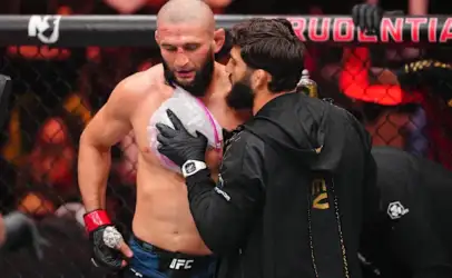 UFC fighter shirtless in the octagon talks with a coach wearing a black jacket inside the cage.