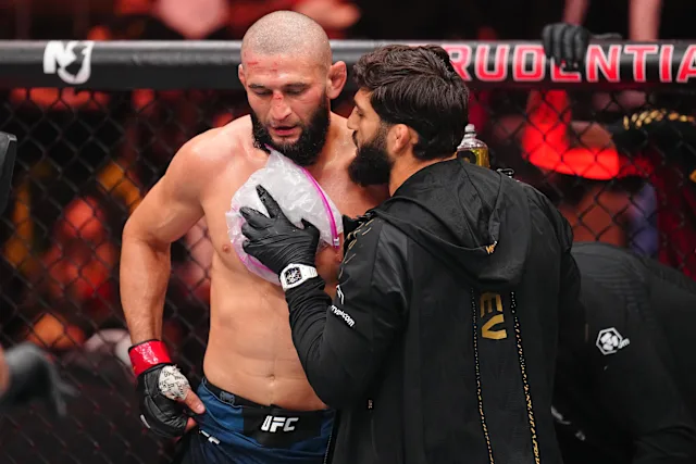 UFC fighter shirtless in the octagon talks with a coach wearing a black jacket inside the cage.