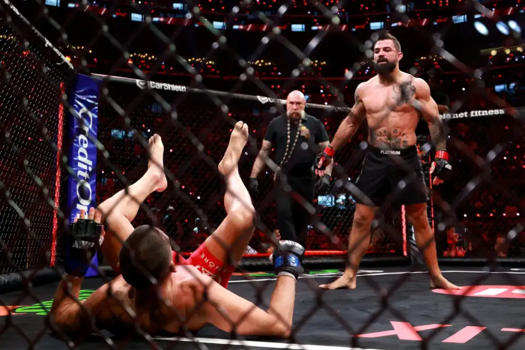 Muscular MMA fighter stands inside the fenced octagon as an opponent lies on the mat with legs up, referee watching in the background.