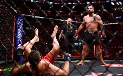 Muscular MMA fighter stands inside the fenced octagon as an opponent lies on the mat with legs up, referee watching in the background.